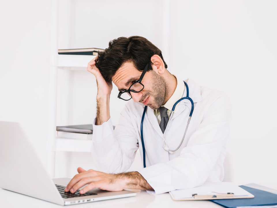 Médico con bata y estetoscopio muestra signos de agotamiento frente a su computadora. Representa el estrés laboral en el sector salud.